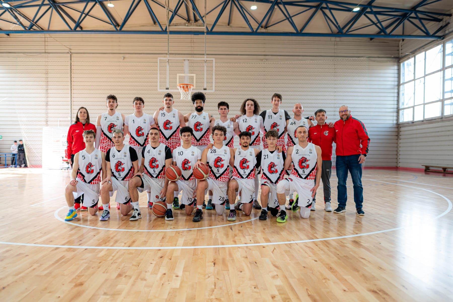 Basketball team in white uniforms and red jackets posing for a group photo on court.