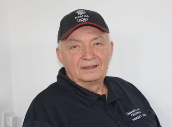 Older man wearing a black Team GB cap and a black polo shirt smiling.