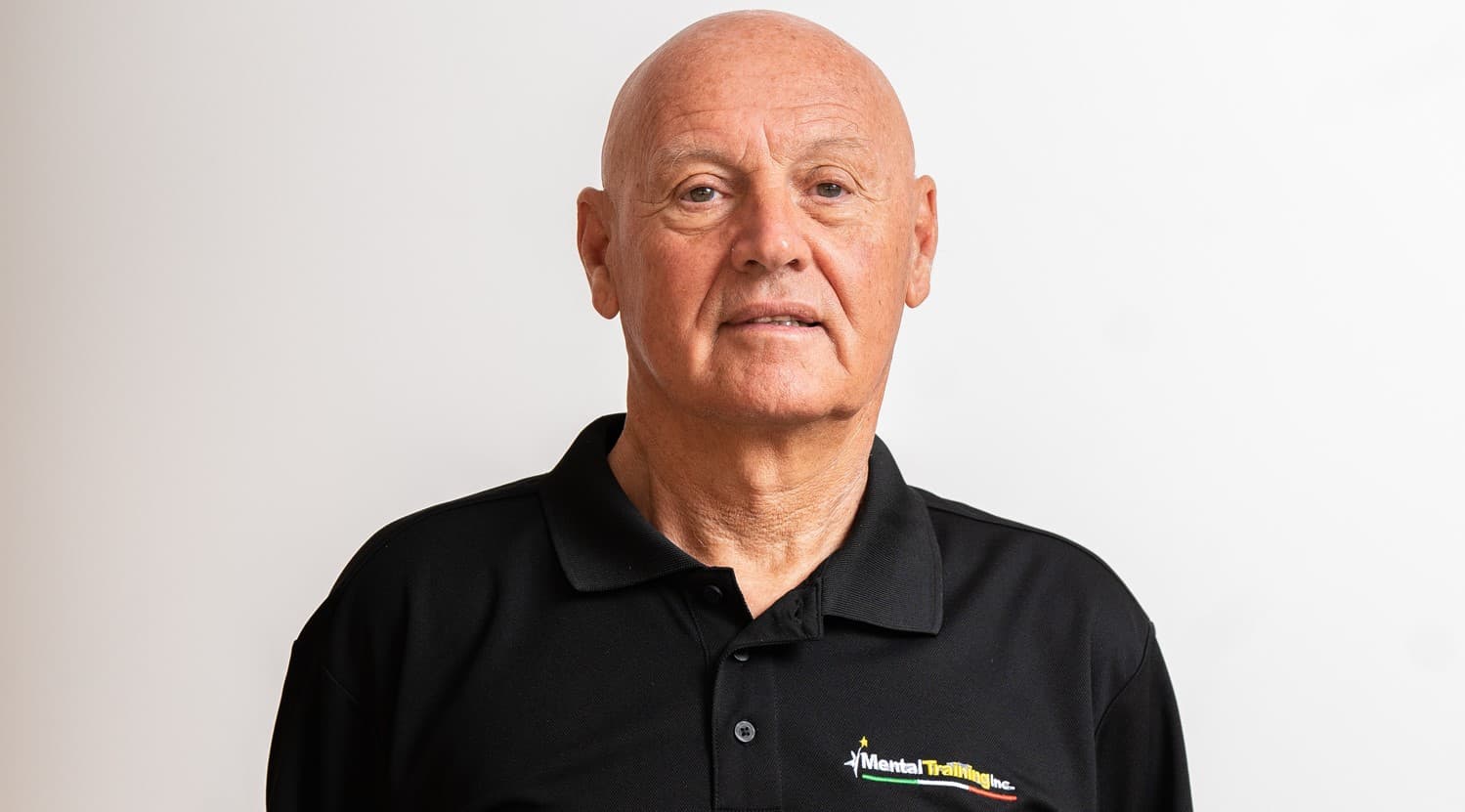 Bald older man in a black polo shirt with a logo against a white background.