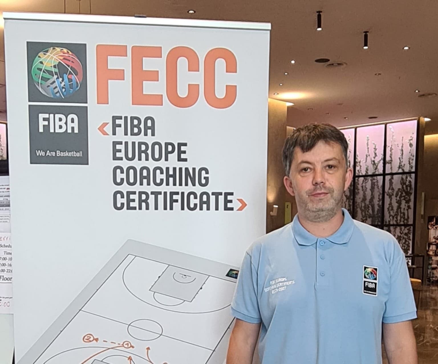 Man in a blue polo shirt standing beside a FIBA Europe Coaching Certificate banner.