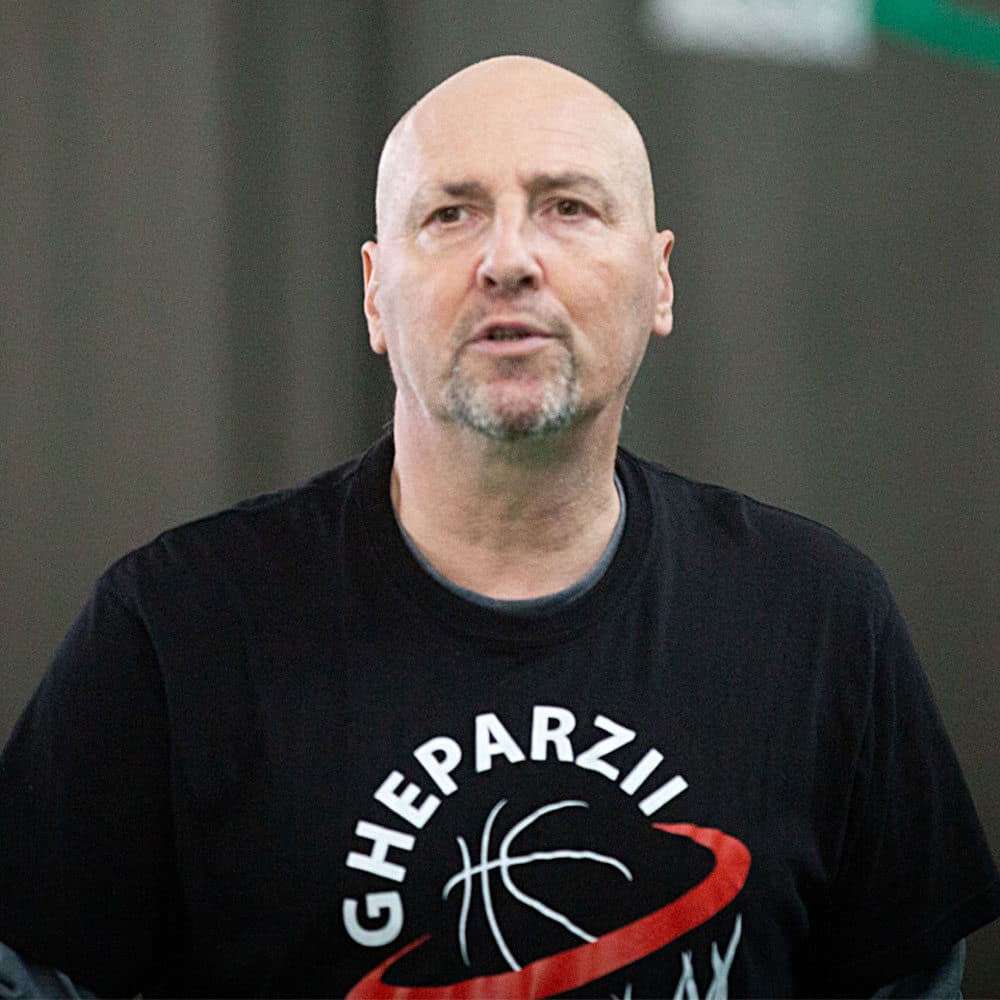 Bald man with a goatee wearing a black GHEPARZII basketball t-shirt looking off-camera.